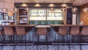 A bar area with a long counter, multiple bar stools, and a display of bottles. - Hotel Libertas elements pure (Merzhausen)