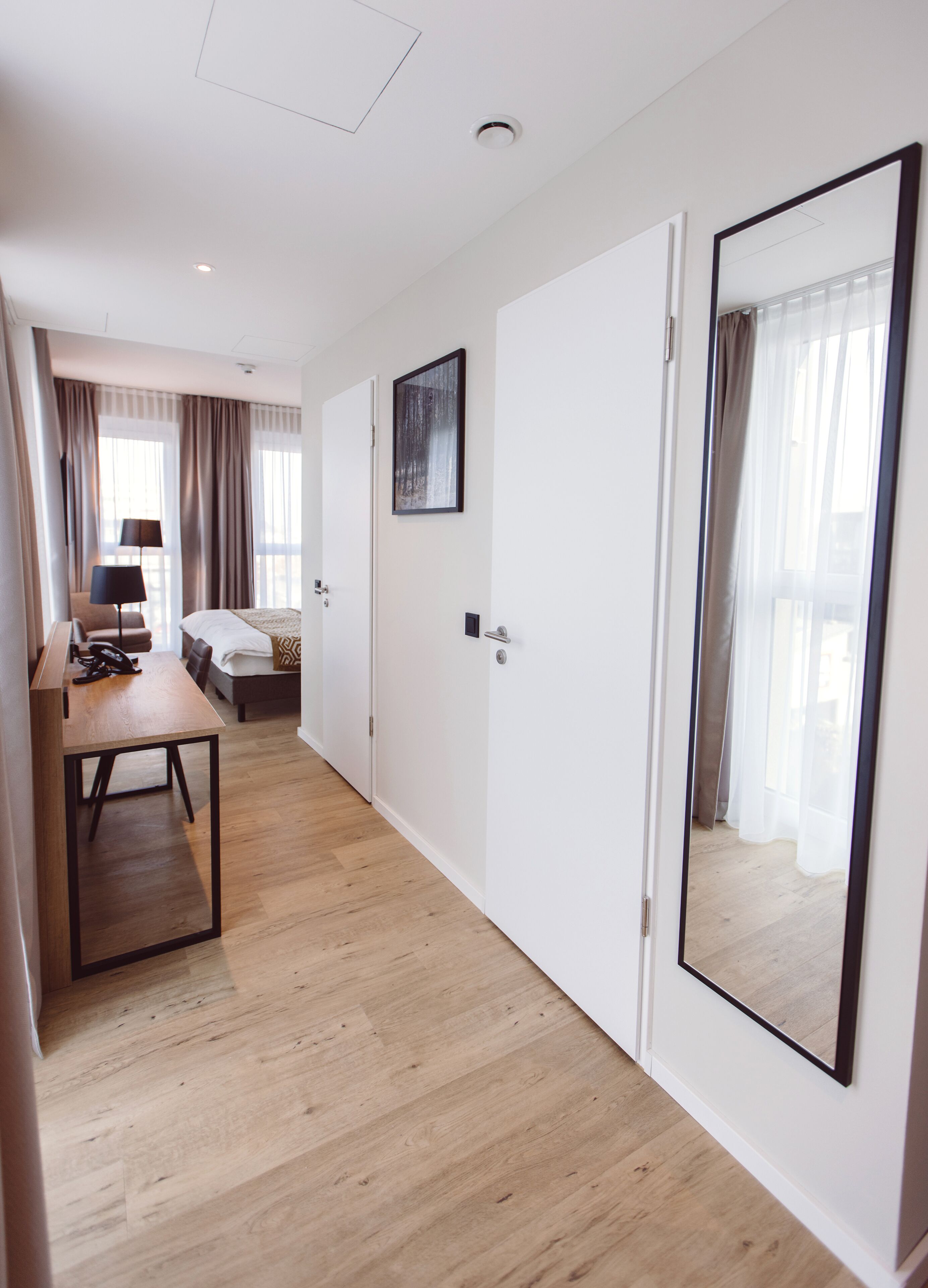 A hotel room with a wooden floor, a bed, a desk with a chair, a lamp, and a large mirror.