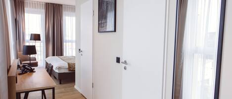A hotel room with a wooden floor, a bed, a desk with a chair, a lamp, and a large mirror.