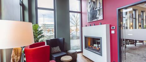 A modern lounge area with a fireplace, red and grey seating, large windows, and a city view.