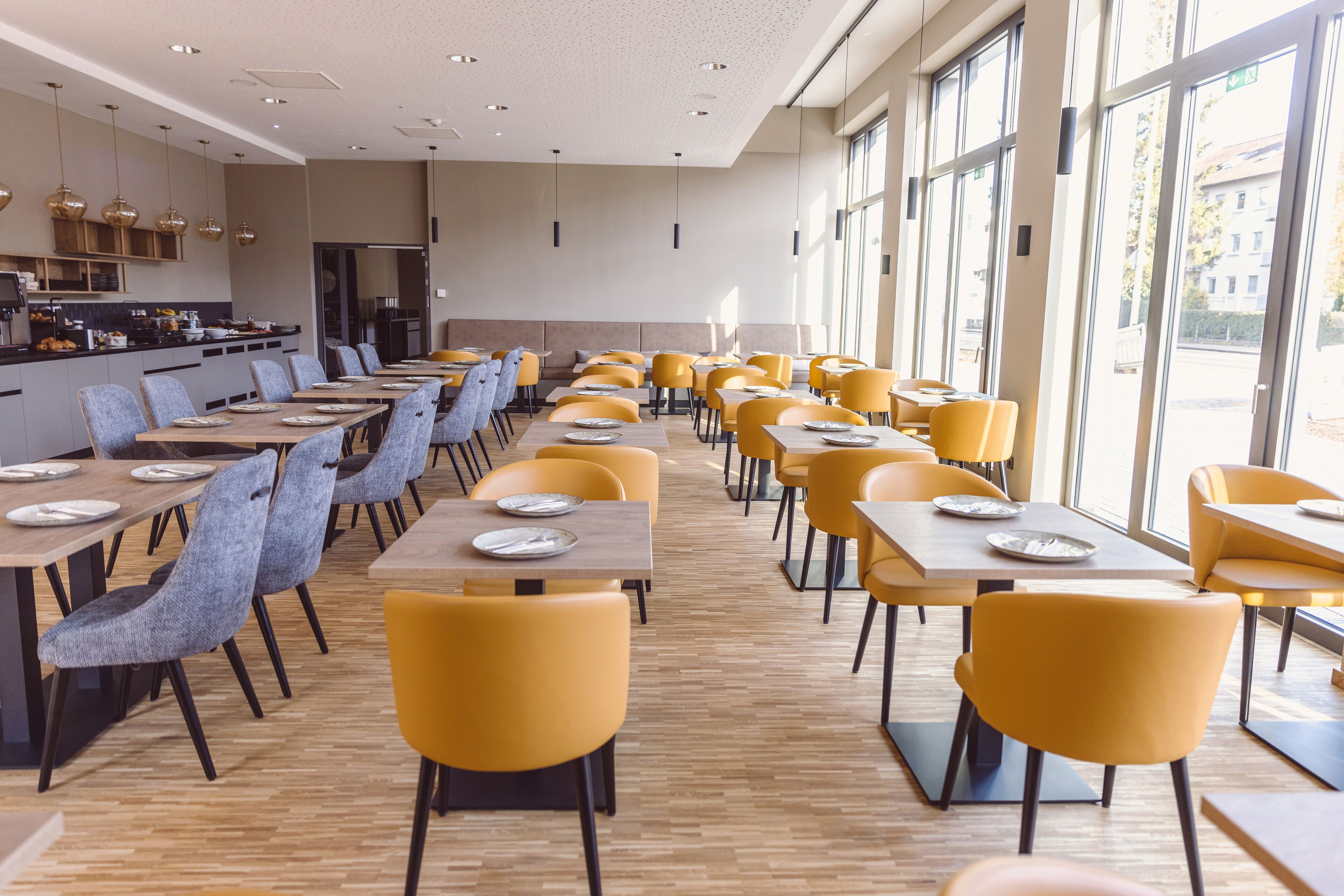 A spacious dining area with multiple tables and chairs, large windows providing natural light, and a modern kitchen in the background.