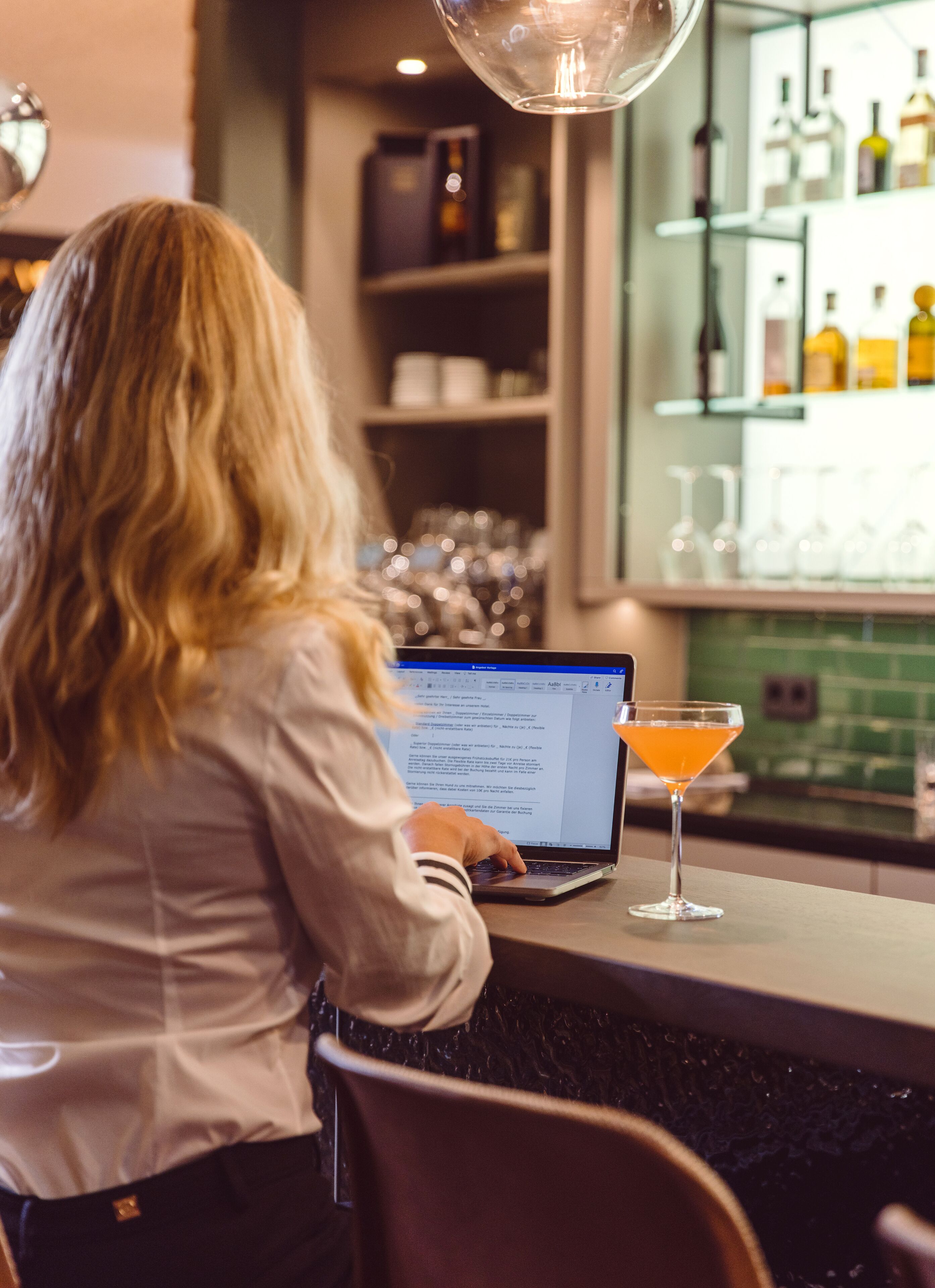 A person using a laptop at a bar with a cocktail on the counter.
