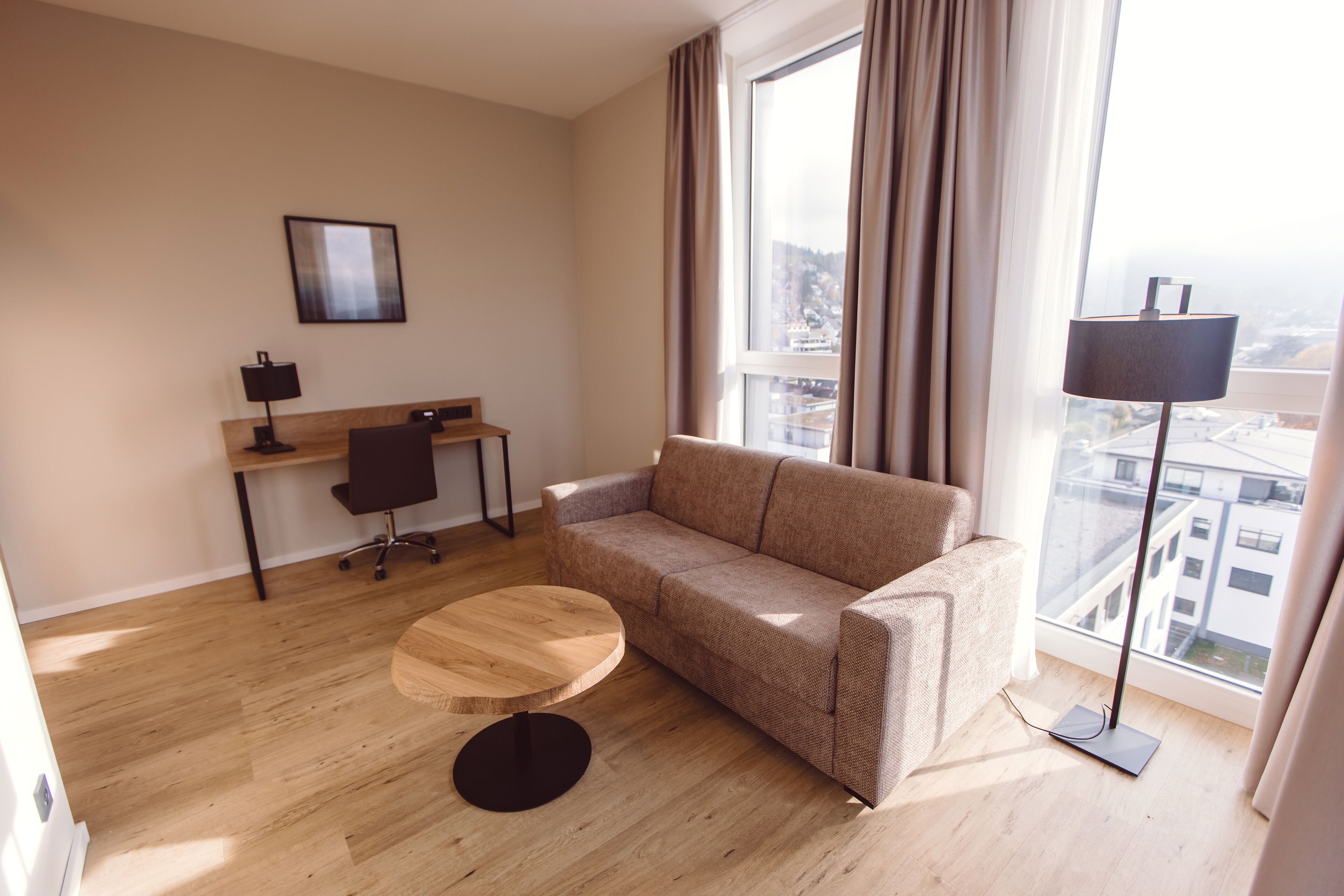 Habitación de hotel moderna con sofá, una mesa redonda de madera, un escritorio con silla y una lámpara de pie.