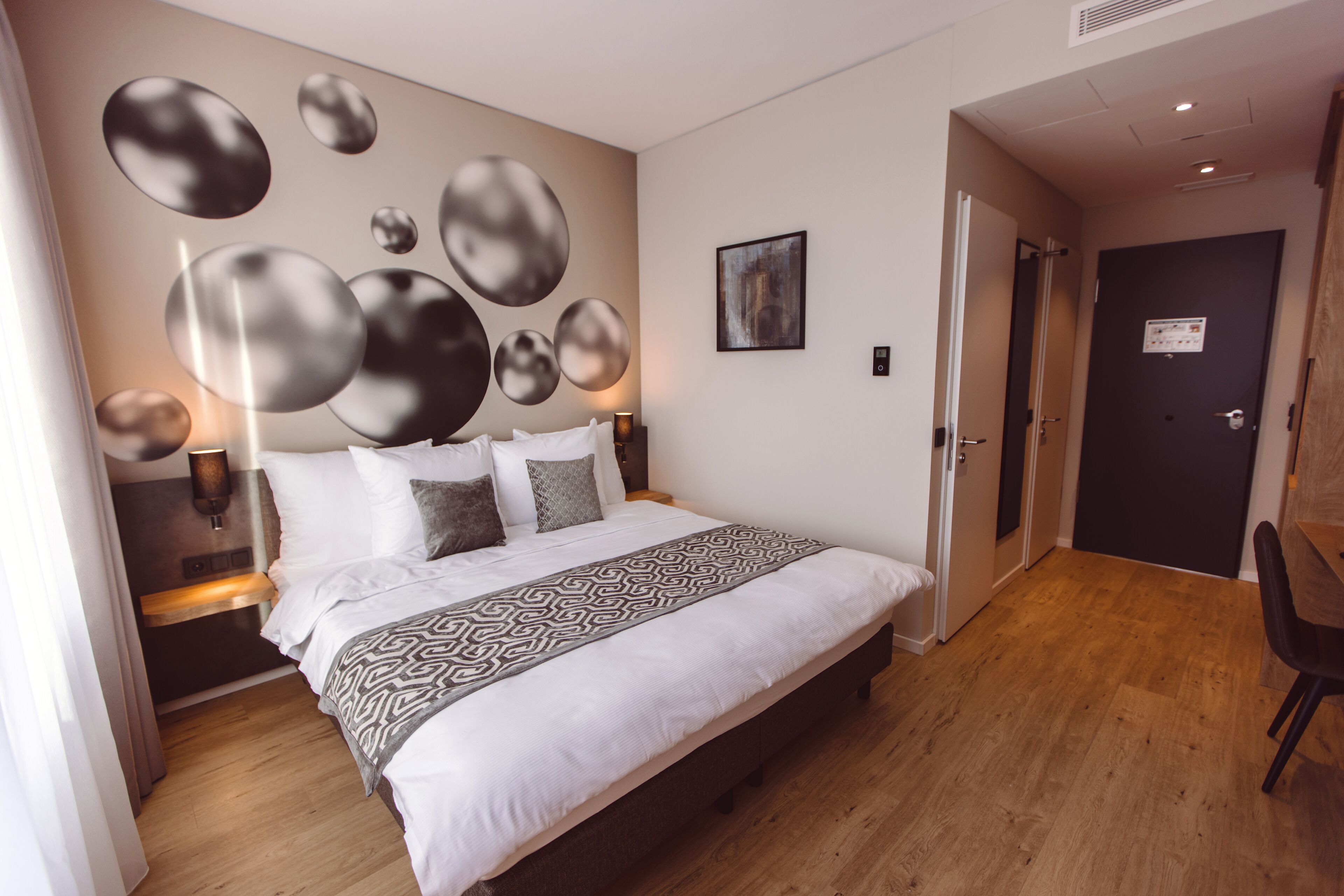 A modern hotel room with a large bed, a wall-mounted abstract art piece, and a door leading to another room.
