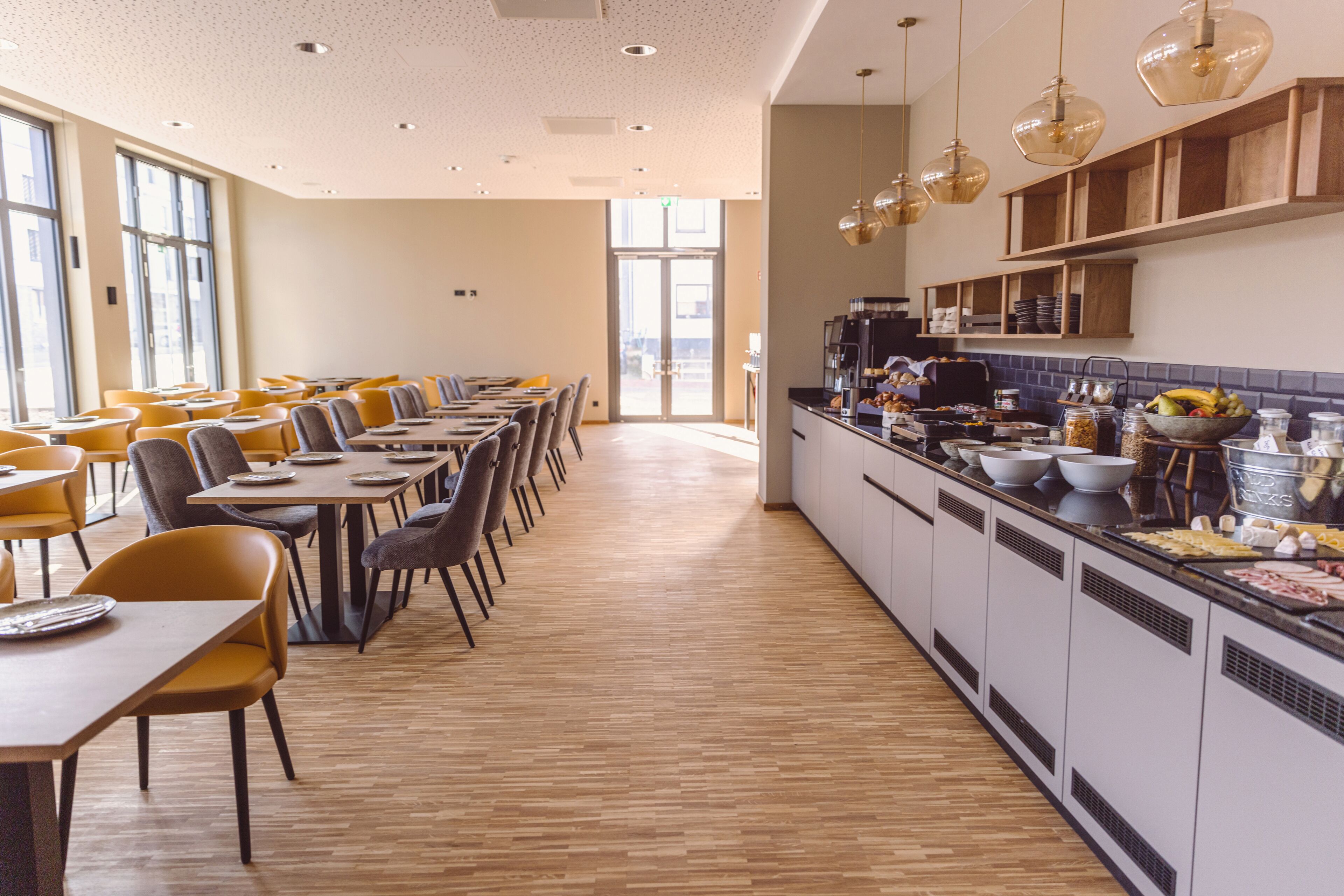 A modern dining area with long tables and chairs, a kitchen counter with food items, and a bright interior with natural light.