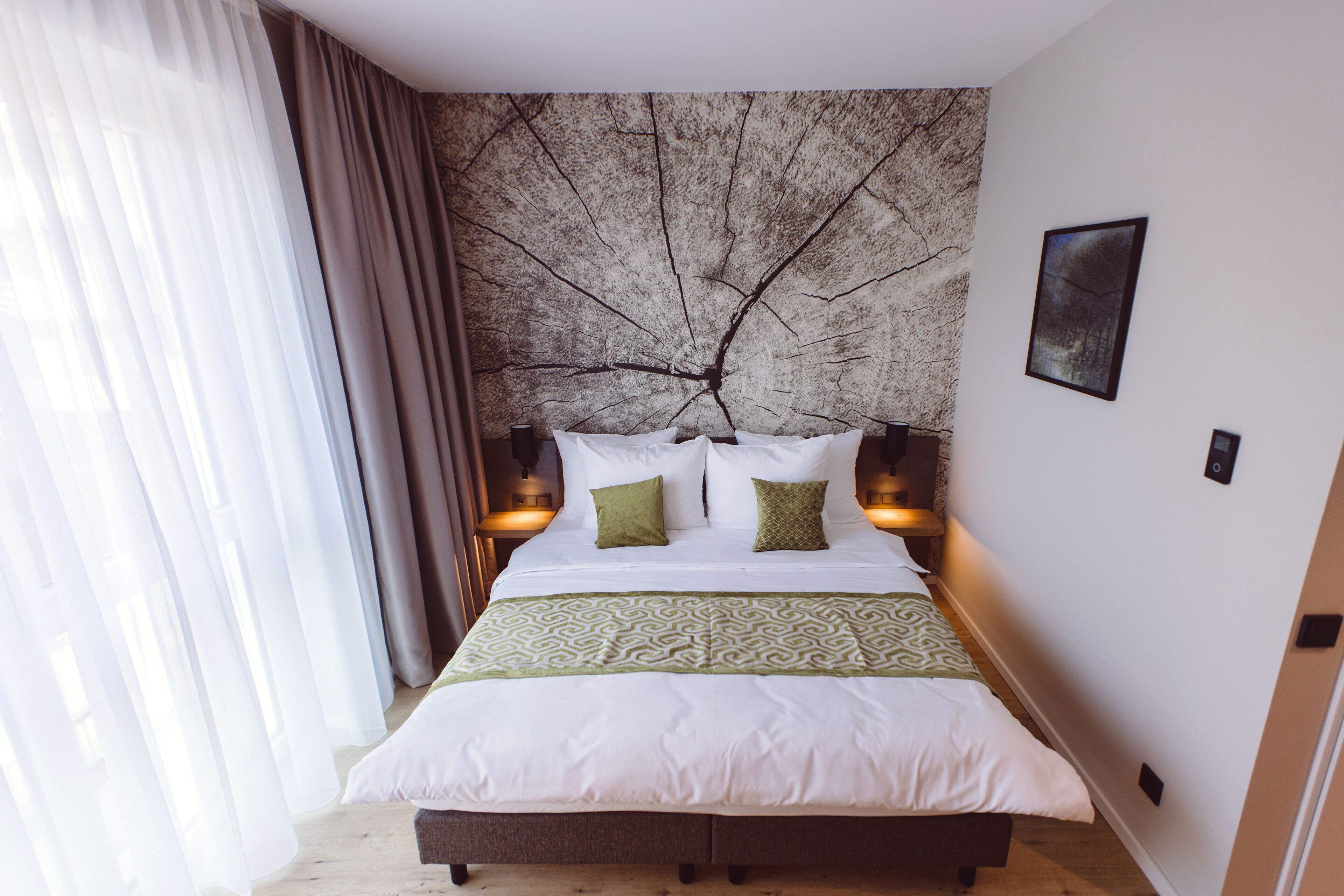 A neatly made bed with white and green bedding, a tree branch mural on the wall, and two bedside lamps.