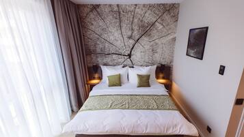 A neatly made bed with white and green bedding, a tree branch mural on the wall, and two bedside lamps.
