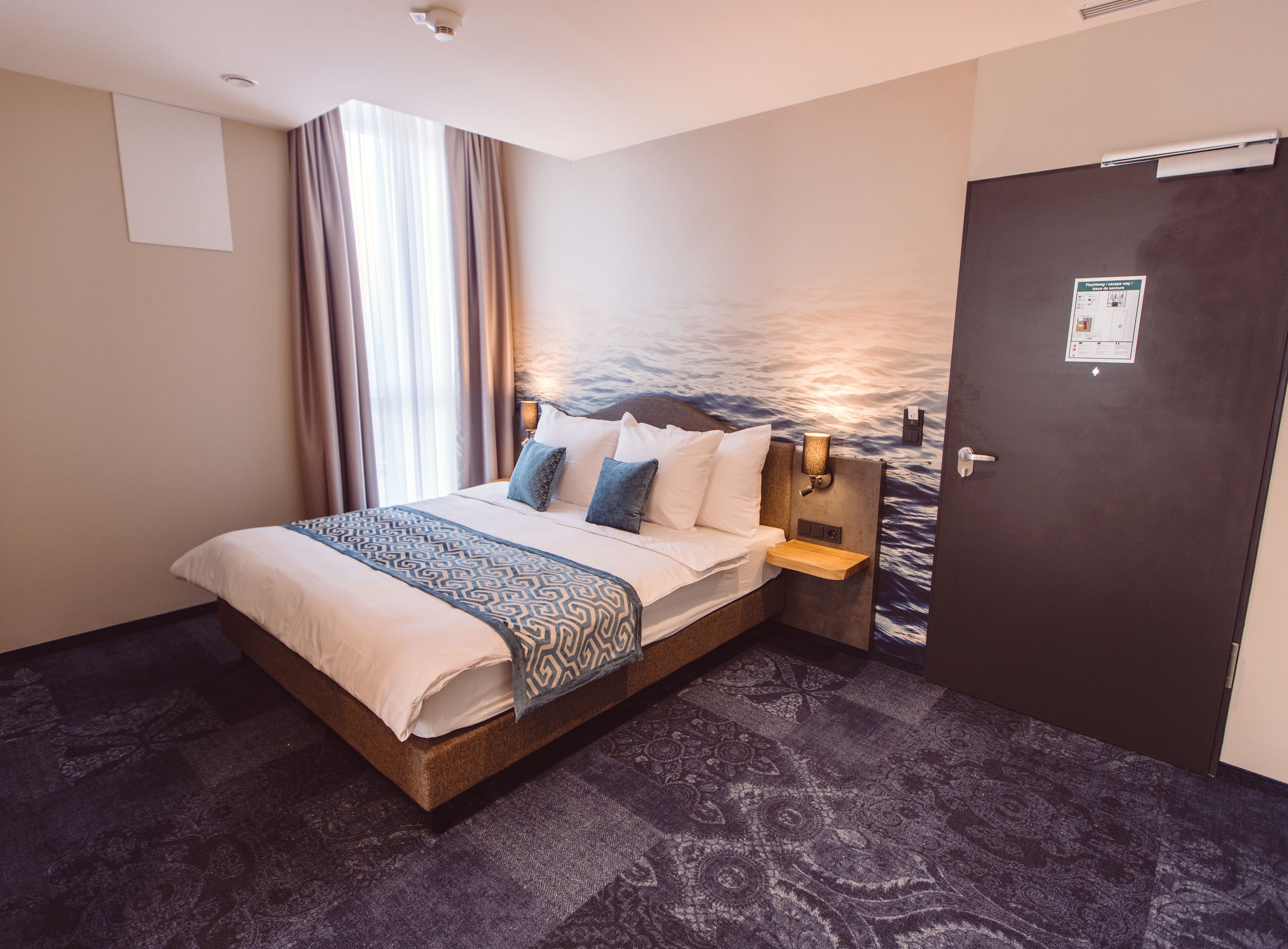 A hotel room with a bed, a nightstand, a wall-mounted lamp, and a door with a sign.
