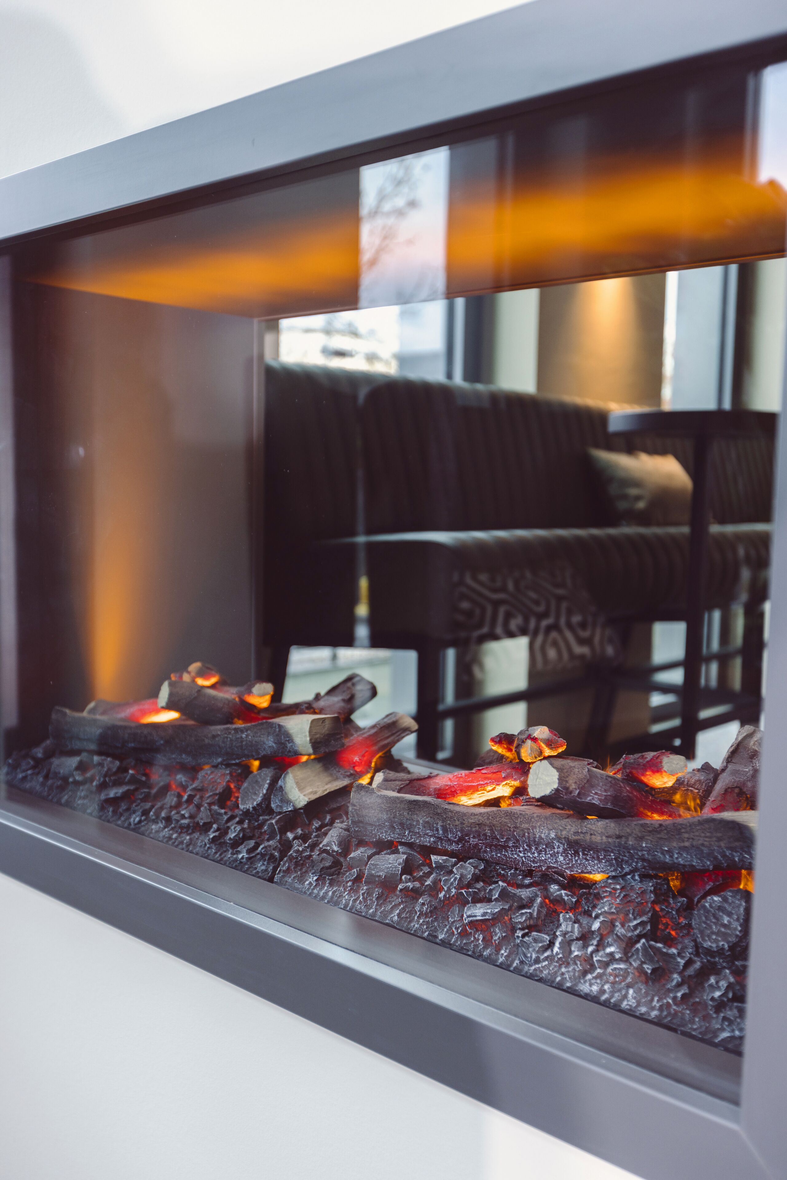 Una chimenea con leña y brasas, un sofá estampado y una ventana que refleja una habitación con una silla.