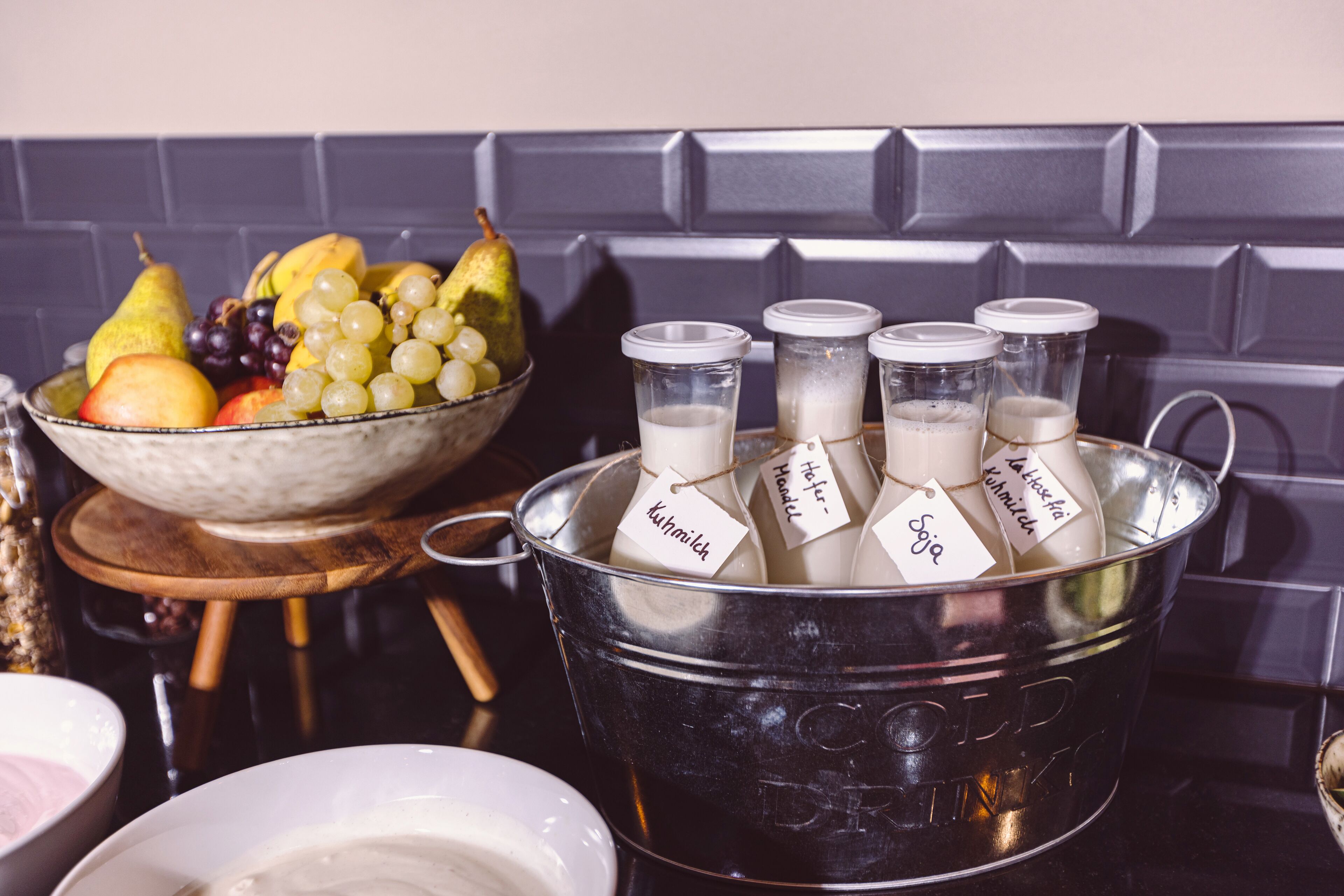 Un balde metálico con botellas etiquetadas con nombres, un cuenco de fruta y un plato blanco.