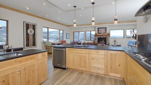 A spacious kitchen with wooden cabinets, a built-in dishwasher, and a large island with a sink. There are pendant lights above the island and a fireplace in the background. - Top-Floor Ski-In/Out Condo with Fireplace, Mountain Views & Peloton Access.Close (Big Sky)