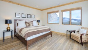 A bedroom with a large bed, two bedside tables, a chair, and a view of a snowy landscape through two windows. - Top-Floor Ski-In/Out Condo with Fireplace, Mountain Views & Peloton Access.Close (Big Sky)
