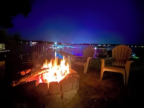 Ein Lagerfeuer mit Sitzgelegenheiten, Blick auf einen Steg mit Lichtern und ein Gewässer.
