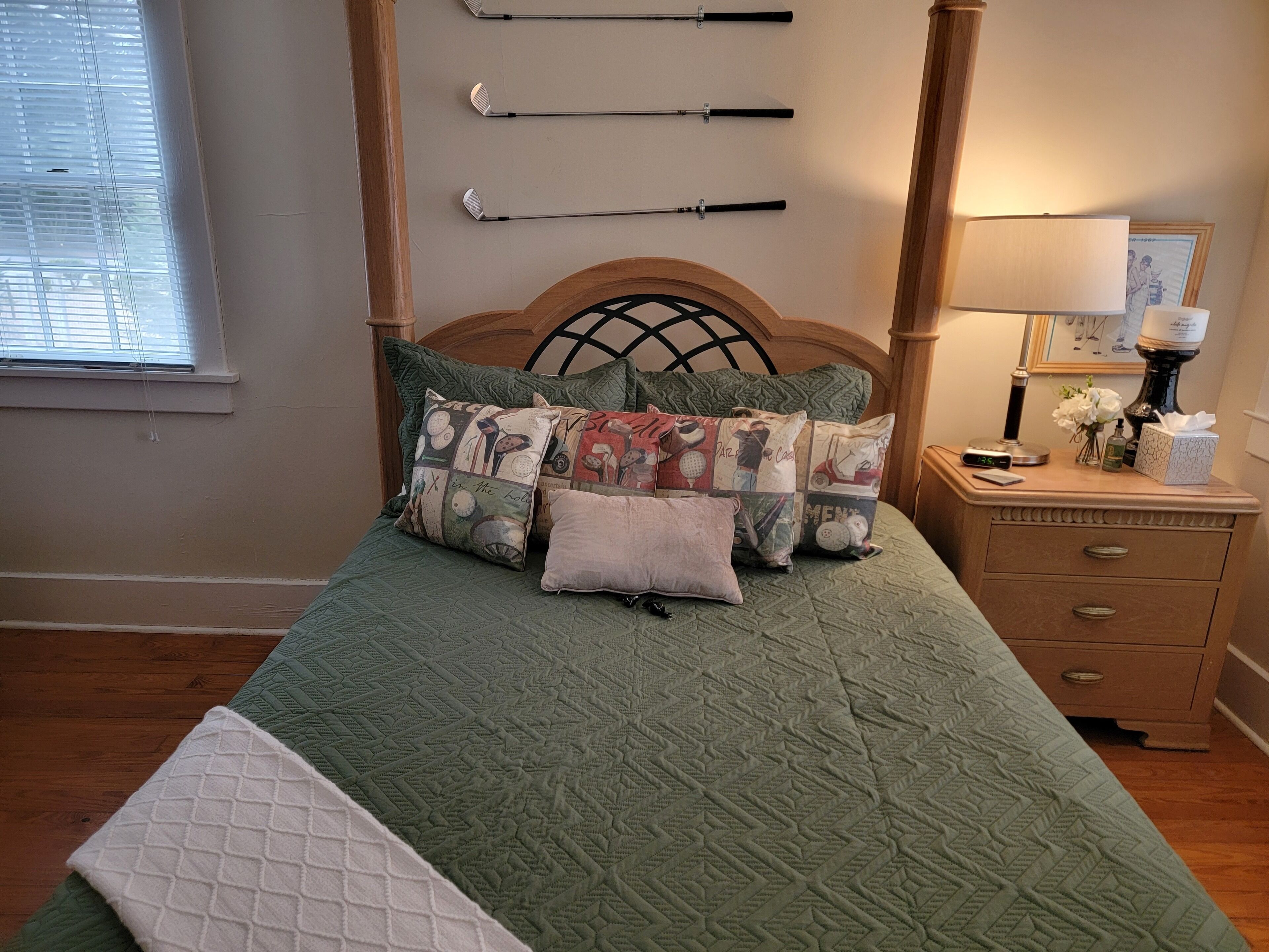 A neatly made bed with a green quilt and multiple decorative pillows, a wooden headboard, a bedside table with a lamp, and a clock.