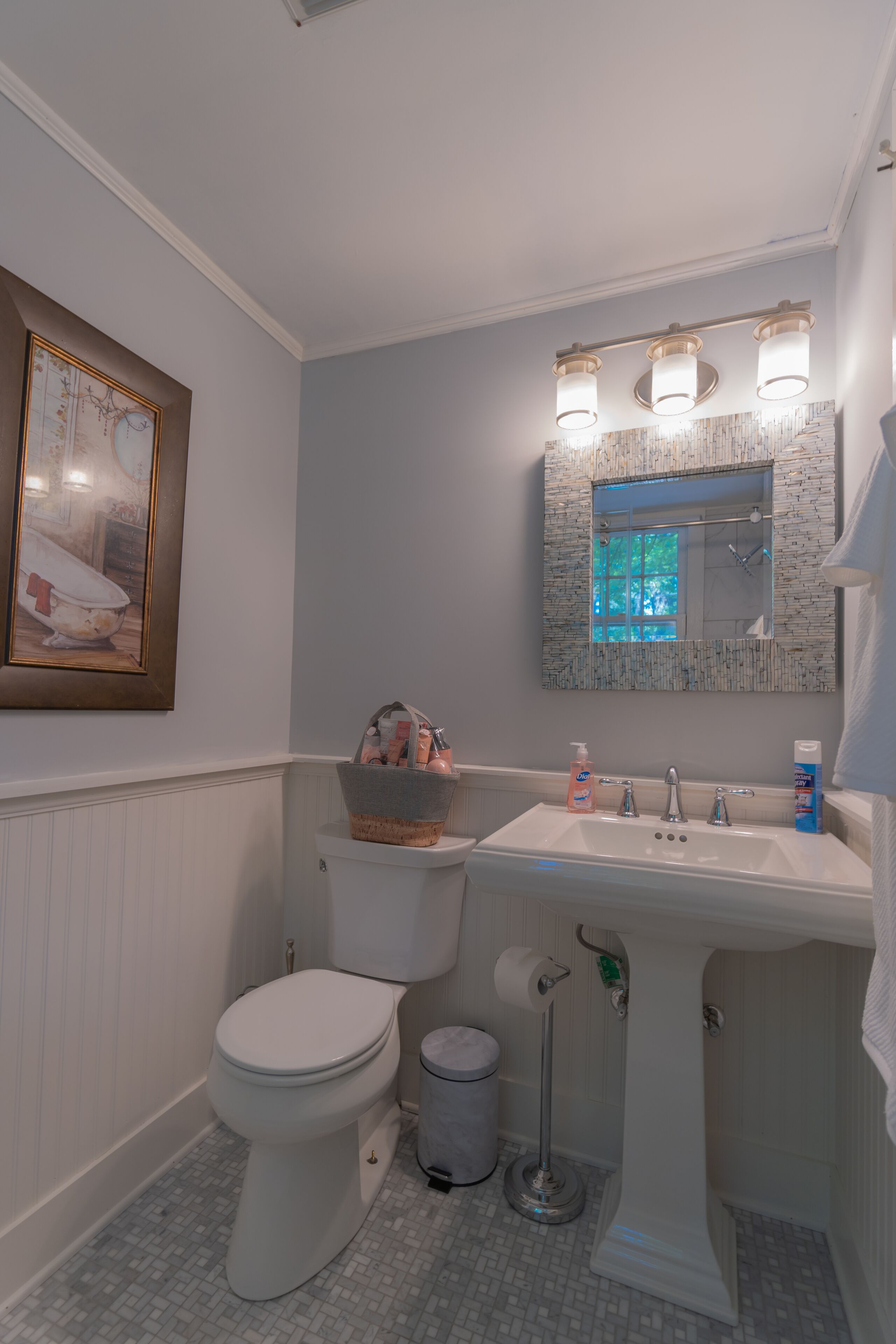Ein Badezimmer mit Toilette, Waschbecken und Spiegel. An der Wand hängt ein gerahmtes Bild und auf dem Waschbecken steht ein Tablett mit Pflegeprodukten.