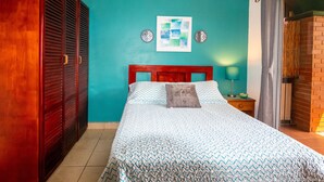 Een slaapkamer met een blauwgroene wand, een houten hoofdbord, een bed met beddengoed met een patroon, twee wandklokken en een nachtkastje met een lamp.