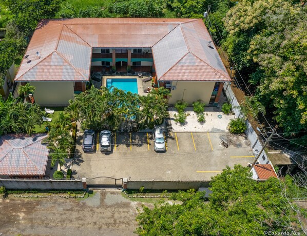 An aerial view of a hotel with a swimming pool, multiple parking spaces, and lush greenery.