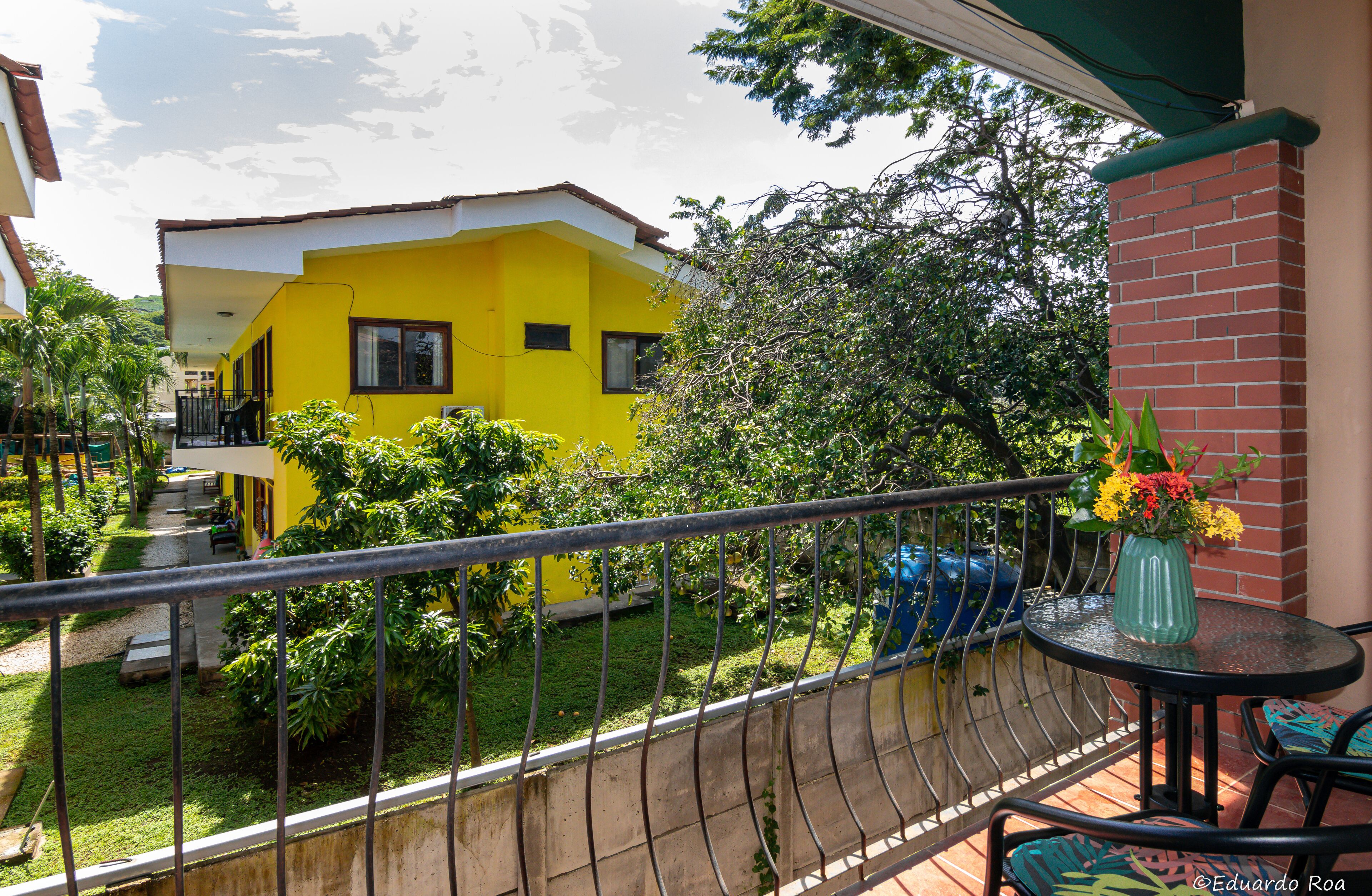 Un edificio amarillo con balcón, un pequeño jardín y una mesa con un florero.