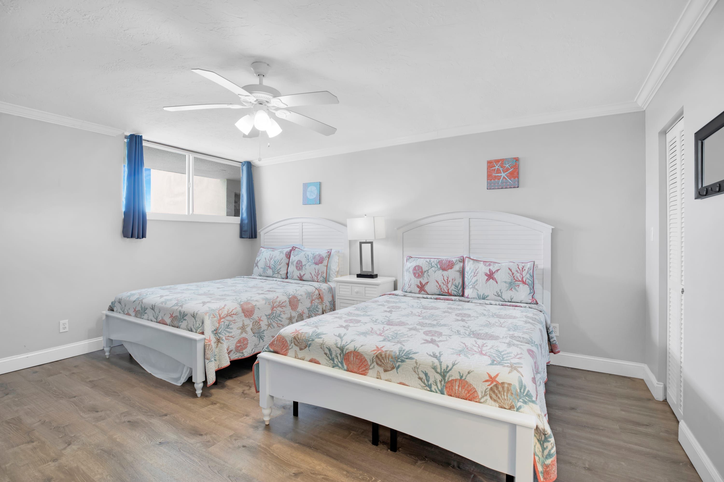 A bedroom with two beds, a ceiling fan, a window with curtains, and a nightstand with a lamp.