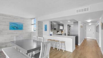 Une cuisine moderne avec des meubles blancs et un îlot central, un coin repas avec une grande table et des chaises blanches, et un couloir menant à une porte.