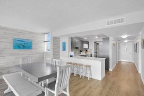 A modern kitchen with white cabinets and a central island, a dining area with a large table and white chairs, and a hallway leading to a door.