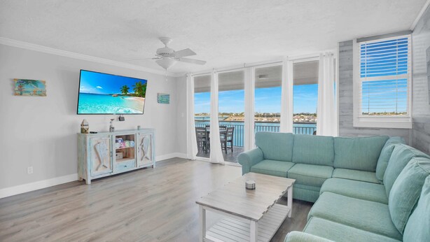 A spacious living room with a large flat-screen TV, a teal sofa, a coffee table, and a view of the ocean through large windows. - Waterfront Dock Pool Condo Near Duval St Pet OK (Key West)