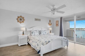 A bedroom with a bed, nightstands, a ceiling fan, and a view of the sea through a sliding glass door.
