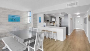 A modern kitchen with white cabinets and a central island, a dining area with a large table and white chairs, and a hallway leading to a door. - Waterfront Dock Pool Condo Near Duval St Pet OK (Key West)