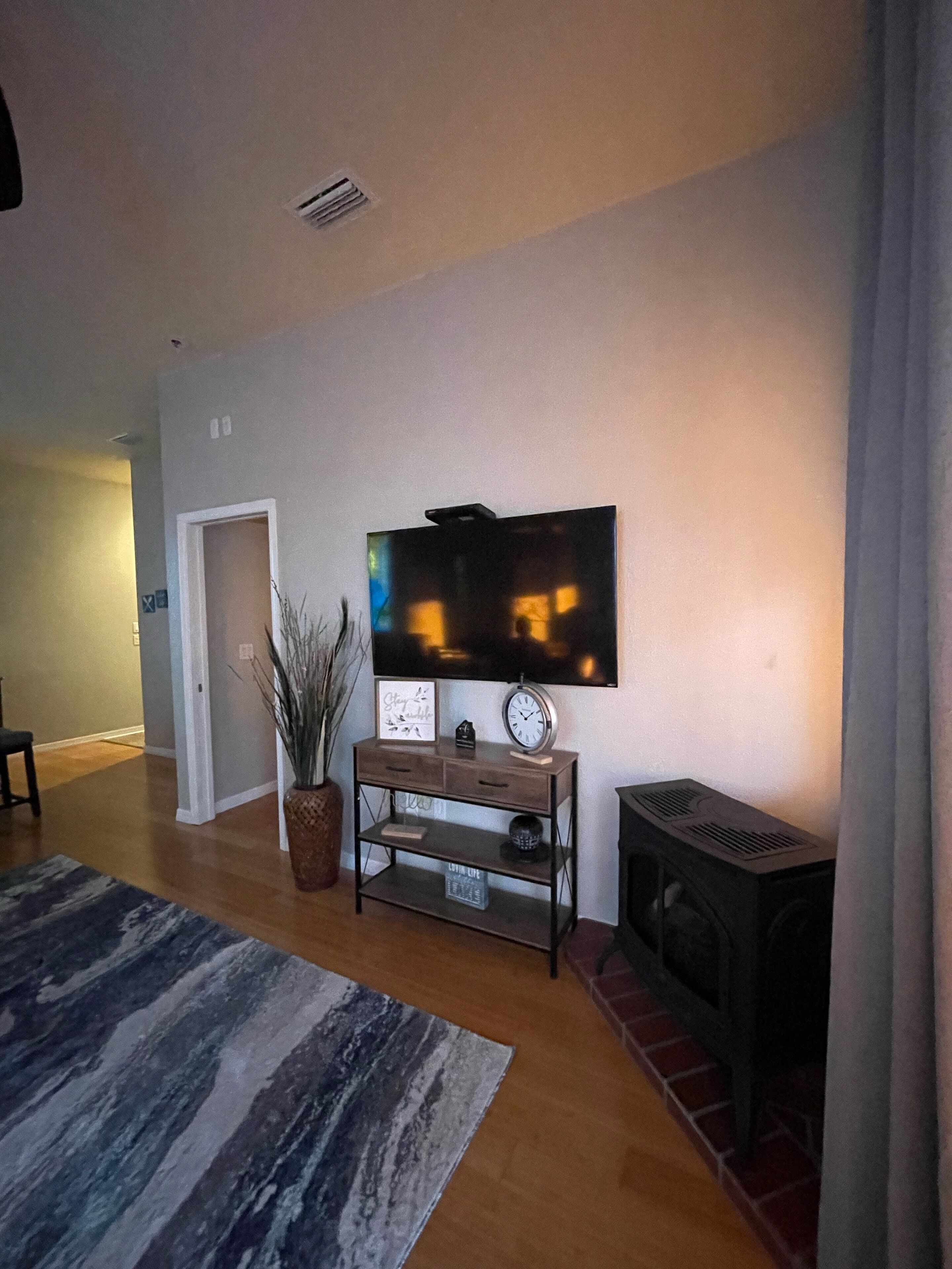 Un salón con televisor de pantalla plana en la pared, una pequeña mesa de madera con un reloj, una planta en maceta y una chimenea.