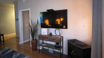 Un salón con televisor de pantalla plana en la pared, una pequeña mesa de madera con un reloj, una planta en maceta y una chimenea.