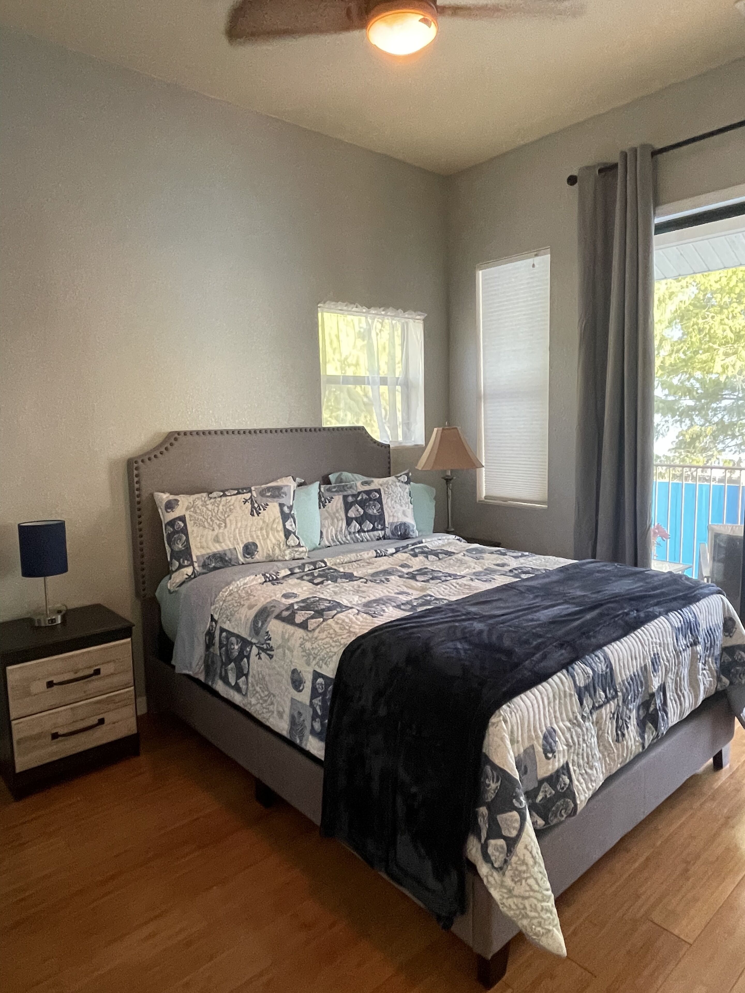 Un dormitorio con cama, mesita de noche, lámpara, ventana y ventilador de techo.