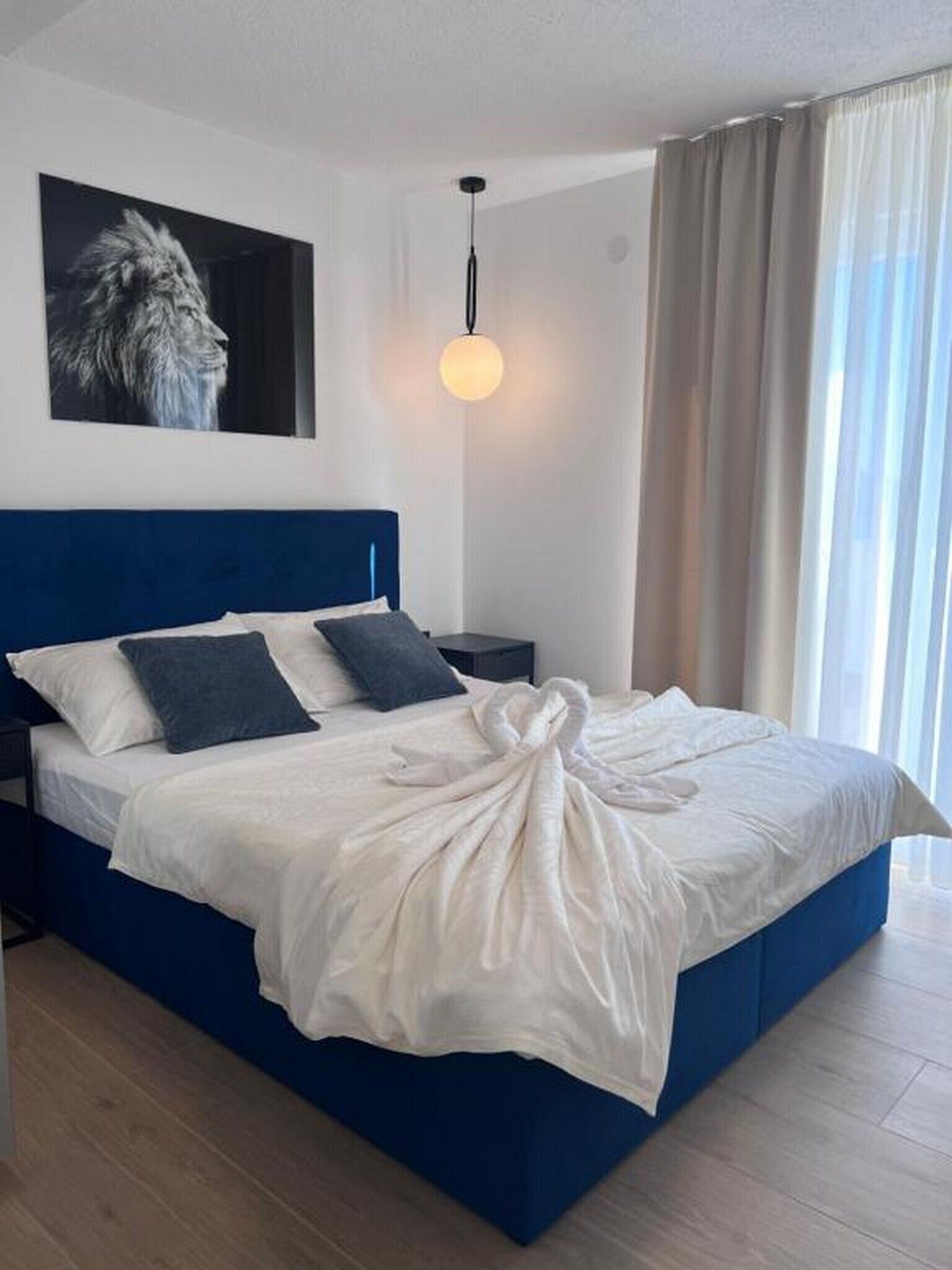Une chambre à coucher moderne avec une tête de lit bleue, un grand lit avec du linge de lit blanc, un plaid, une table de chevet, une lampe et un tableau encadré accroché au mur.