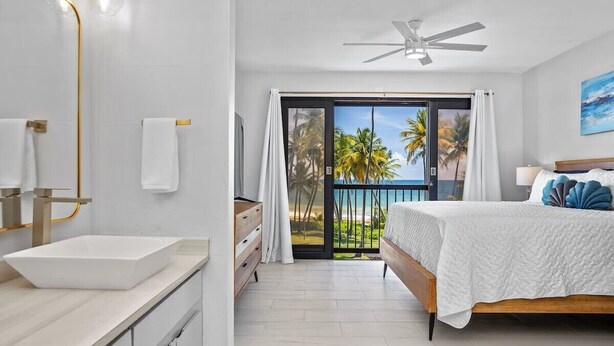 Un dormitorio con cama, un baño con lavamanos y un balcón con vistas a palmeras.