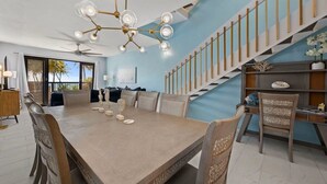 Un comedor amplio con una gran mesa y sillas, una lámpara moderna, una escalera con barandilla de madera y vistas al exterior.