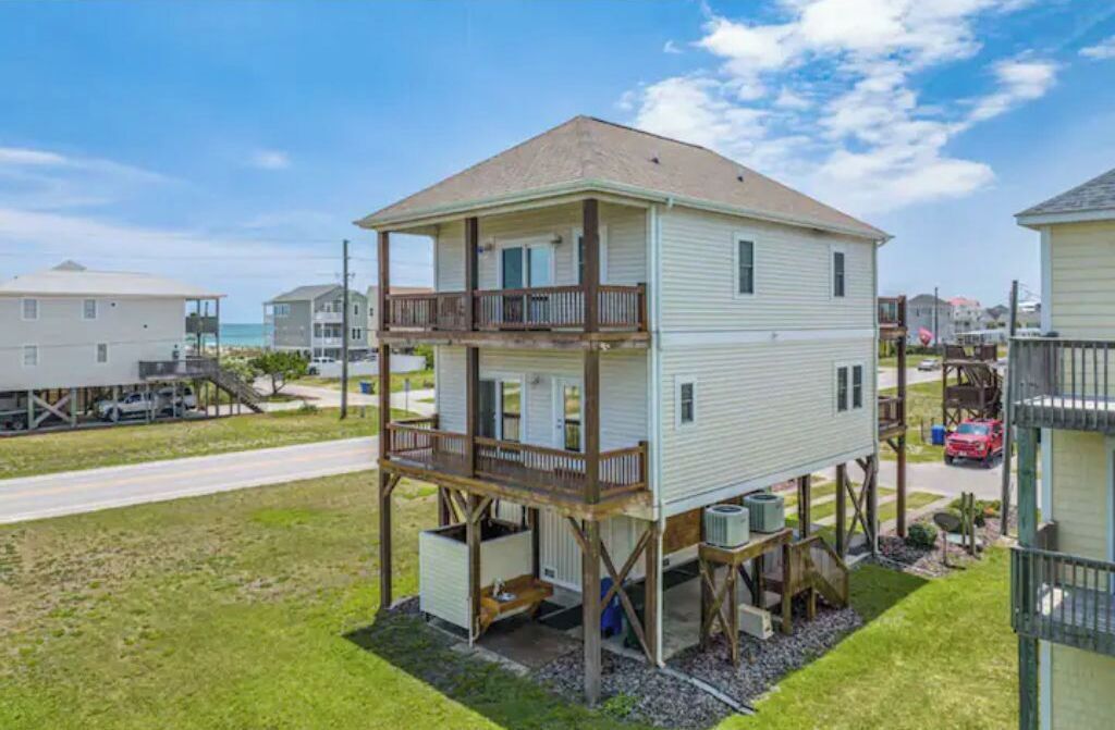 A two-story house with a balcony on the upper floor, elevated on stilts, surrounded by a grassy yard and a road.