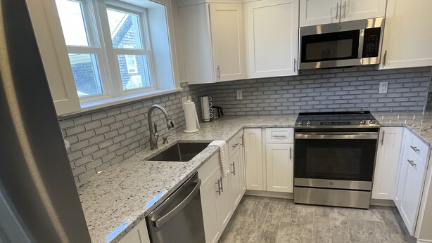 A modern kitchen with white cabinets, a stainless steel refrigerator, a dishwasher, an oven, and a microwave. - Close to everything (Newport)