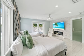 A modern bedroom with two beds, a TV mounted on a brick wall, and a view of the outdoors through a sliding door.