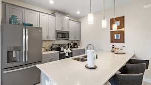 A modern kitchen with grey cabinetry, stainless steel appliances, a central island with seating, and pendant lighting.