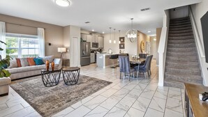 A spacious living area with a sofa, coffee table, and dining table. A kitchen with modern appliances and a staircase leading to an upper level is visible.