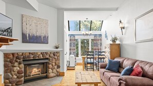 A cozy living room with a stone fireplace, a wooden coffee table, a brown sofa with colorful cushions, and a dining area with a wooden table and chairs. - Lodge at Mtn Village B156 | Ski-In/Out | Valley View | Pool | Hot Tub (Park City)