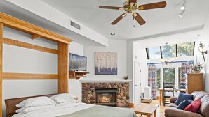 A bedroom with a bed, a fireplace, a ceiling fan, and a view of the outdoors through a large window. - Lodge at Mtn Village B156 | Ski-In/Out | Valley View | Pool | Hot Tub (Park City)