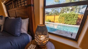 Una camera da letto con testiera in legno, letto con biancheria azzurra, un tavolino rotondo in legno con una lampada intrecciata e una finestra vista piscina e verde.