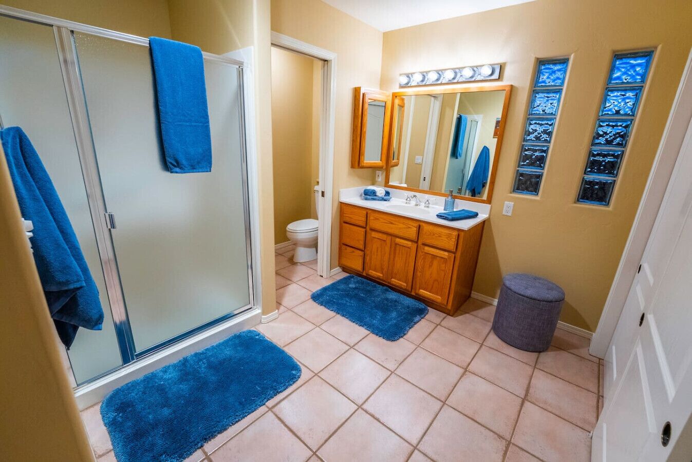 Une salle de bain avec une porte de douche en verre, un meuble-lavabo en bois avec un miroir et un tapis bleu sur le sol.