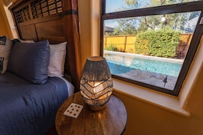 A bedroom with a wooden headboard, a bed with blue bedding, a round wooden table with a woven lamp, and a window with a view of a swimming pool and greenery.