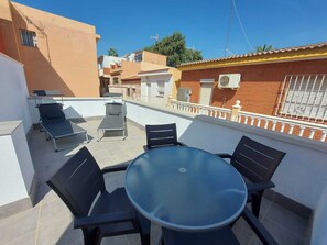 A rooftop terrace with a round table and four chairs, a sunbed, and a view of neighboring buildings.