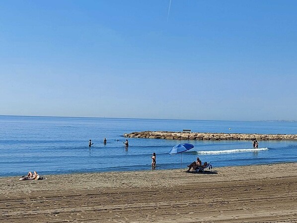 A beach with people swimming and relaxing, a rocky breakwater, and a clear blue sky.