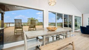 A spacious room with a large wooden table, two chairs, and a bowl. There is a sliding glass door leading to an outdoor deck with a view of the sky and trees.