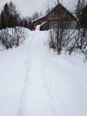 Un sentier enneigé mène à un chalet en bois dont on aperçoit la porte et les fenêtres.