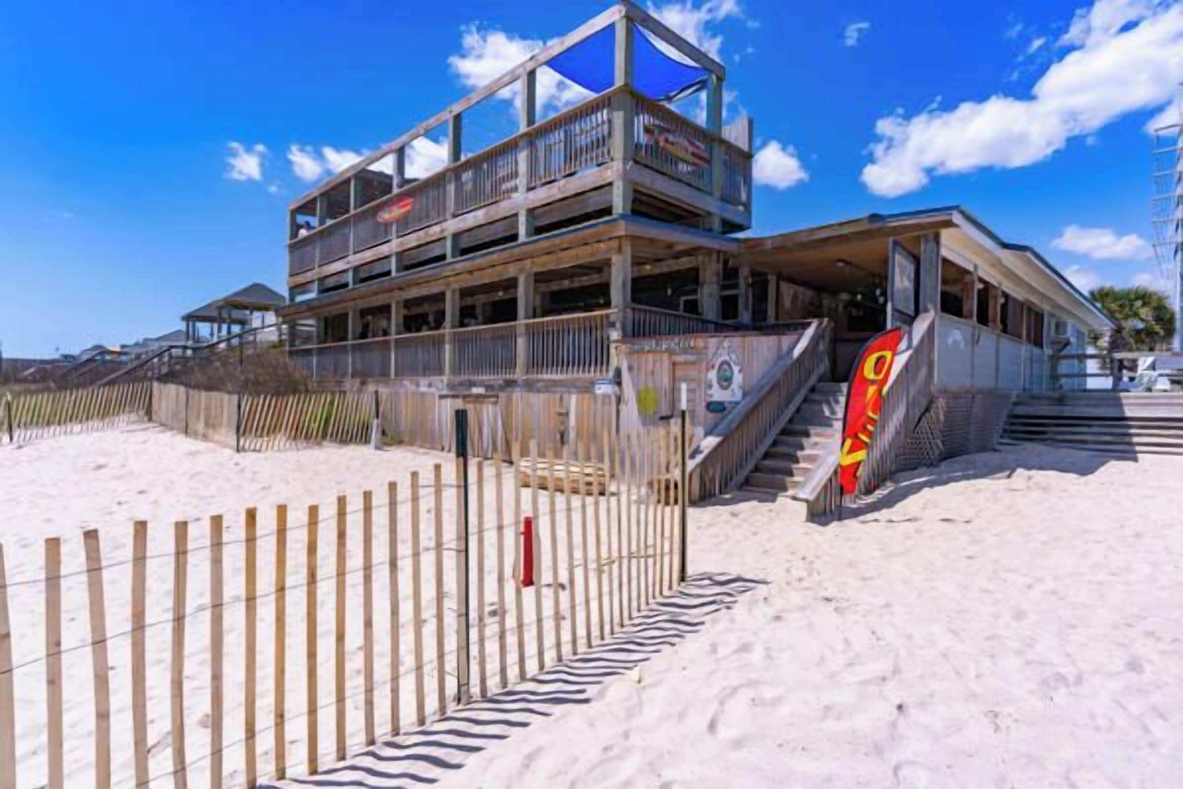 Un edificio de varios pisos con balcones, una playa arenosa al frente y un cielo azul despejado.
