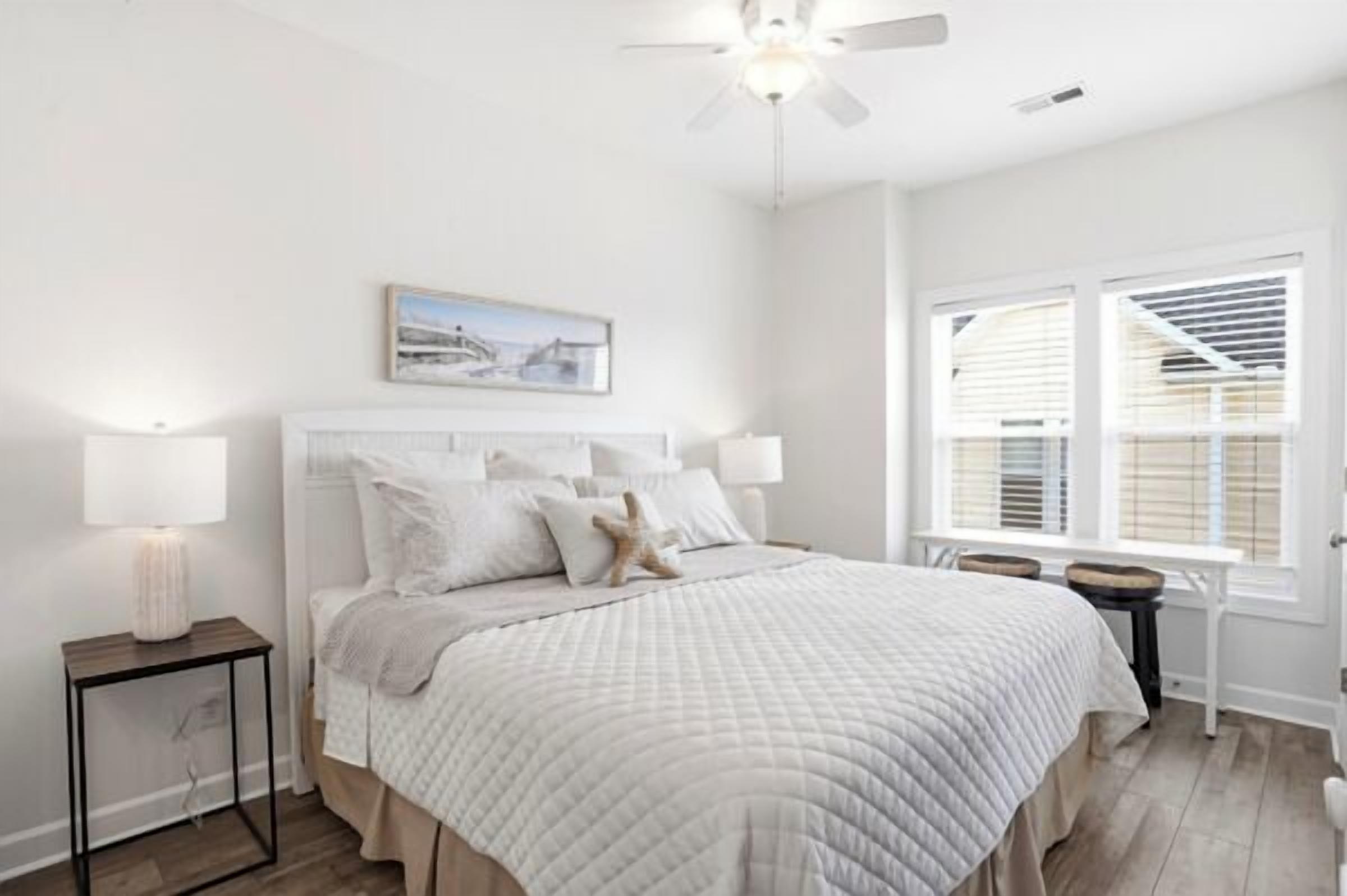 A bedroom with a bed, bedside lamps, a ceiling fan, a window with blinds, and a framed picture on the wall.